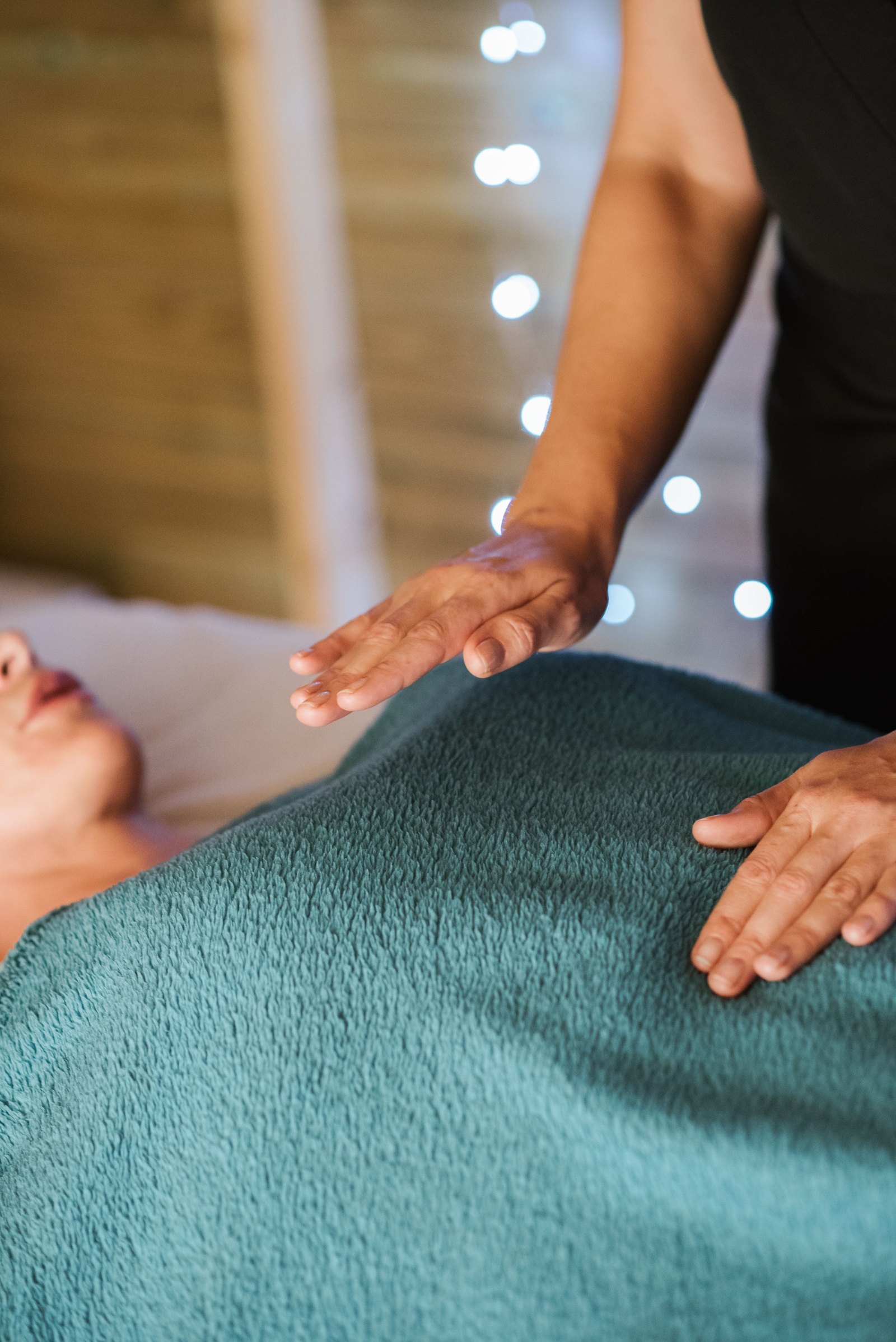 Les mains d&rsquo;une praticienne, l&rsquo;une posée et l&rsquo;autre suspendue au-dessus d&rsquo;une personne allongée sous une couverture sarcelle, une guirlande de lumières en fond