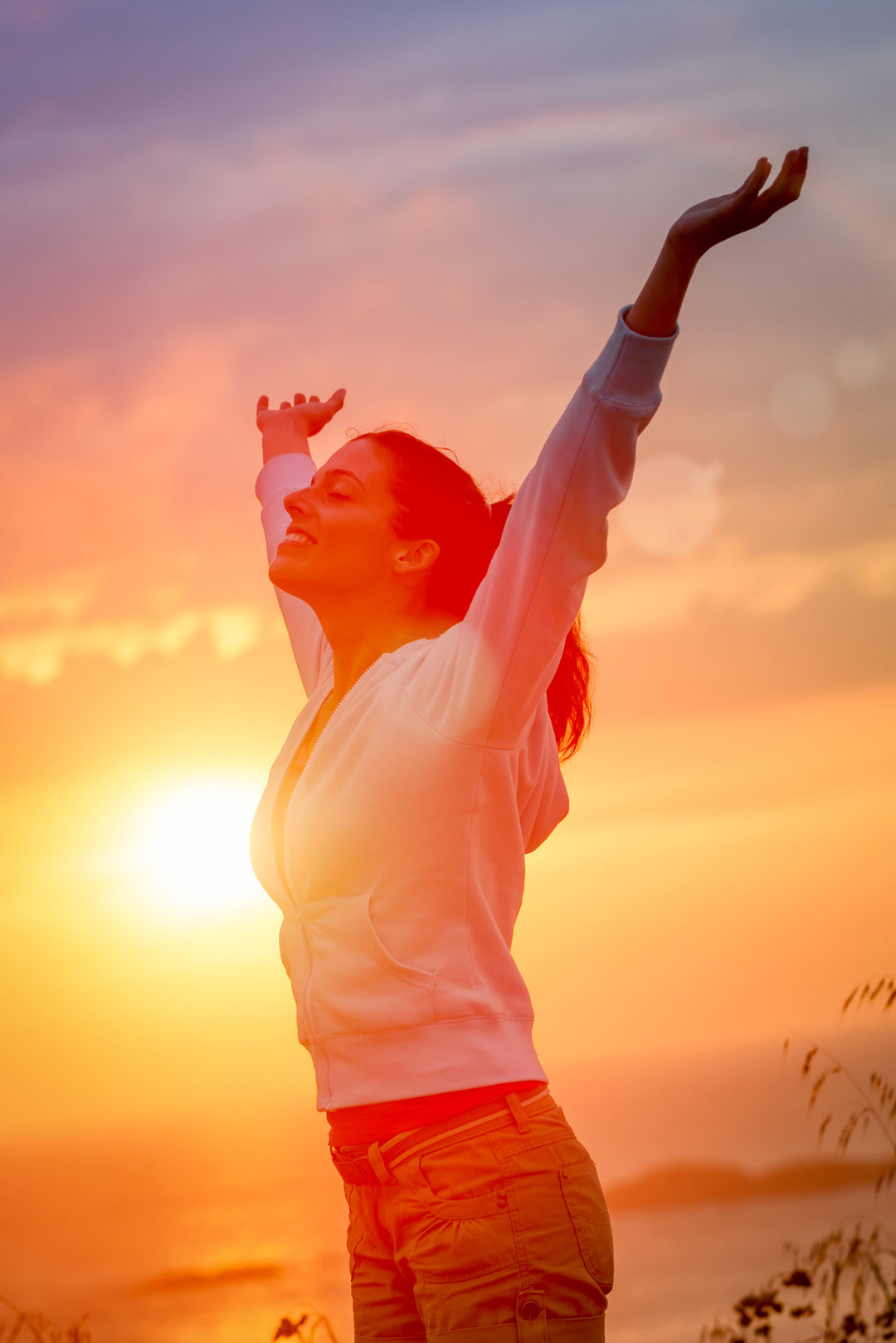 Une femme debout de profil, les yeux fermés et les deux bras levés vers un ciel de coucher de soleil orange et mauve.