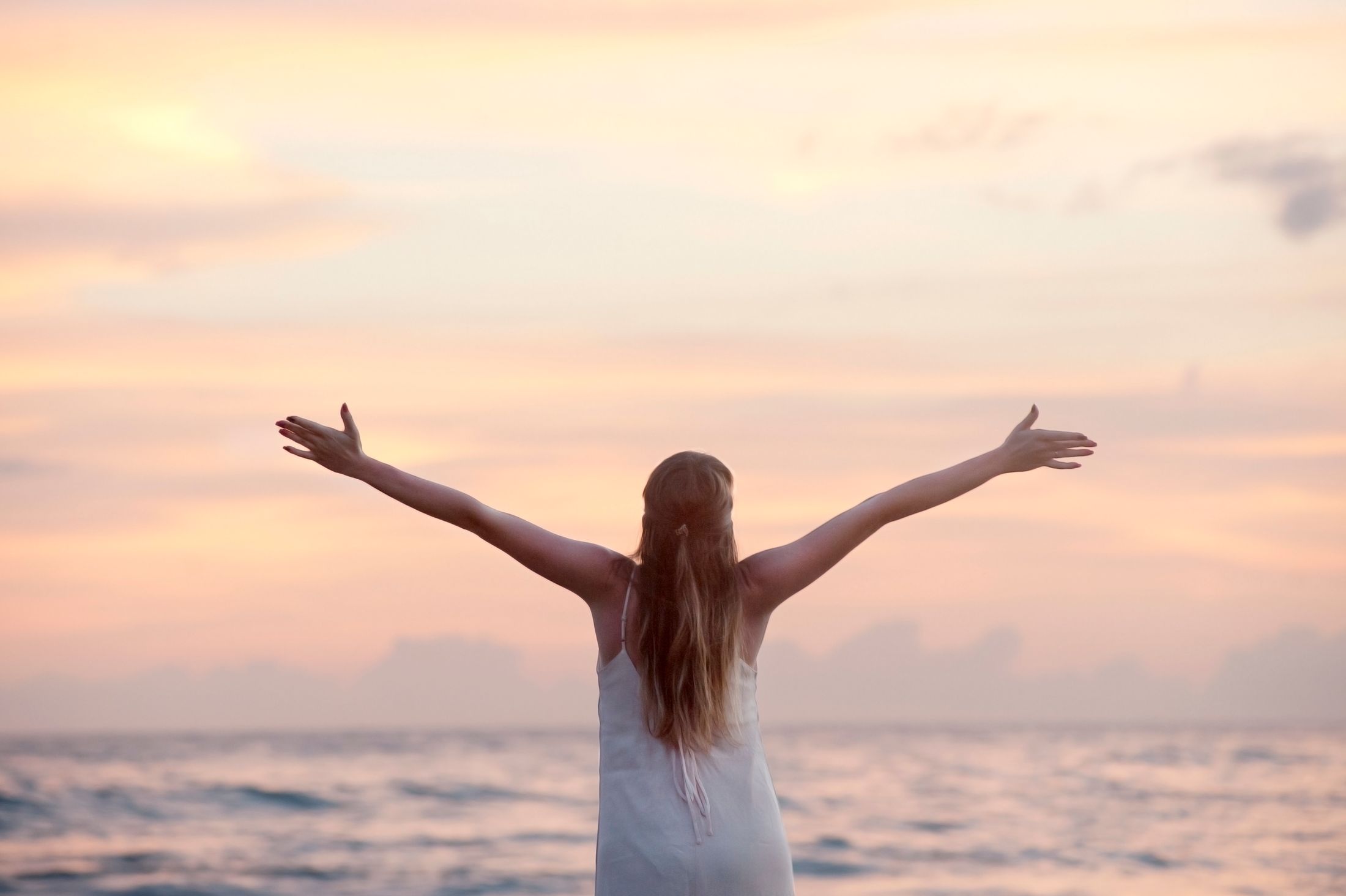 Femme en robe blanche, bras grands ouverts devant la mer sous un ciel pêche