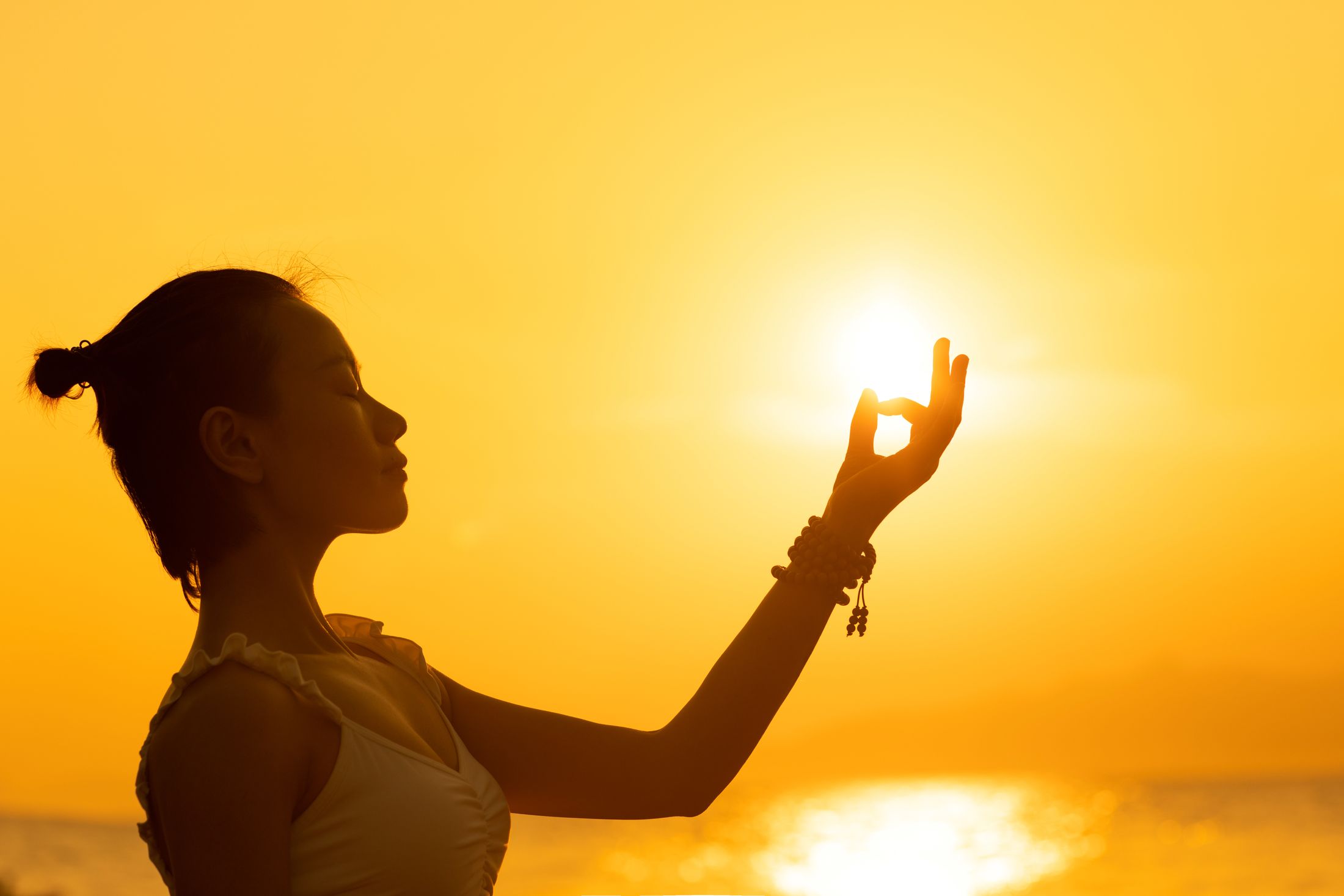 Femme de profil en méditation, la main en mudra devant le soleil, sous un ciel doré